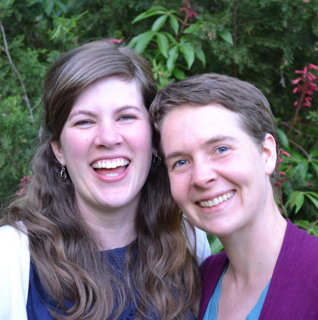Two white women in their 30s, Abby (left) and Sarah (right), are smiling and looking at the camera in front of some greenery